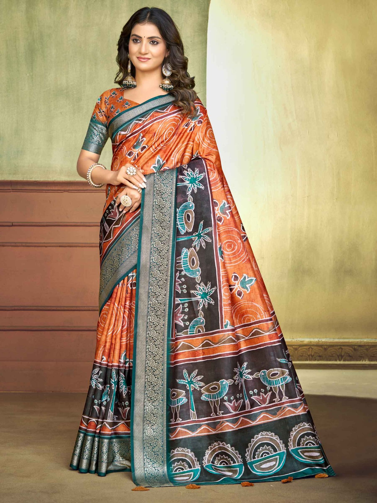 A woman modeling a traditional Indian saree with a vibrant orange and black patterned body, teal borders, and a matching teal blouse, set against a warm beige and brown studio background.